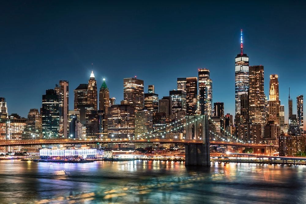 New York City skyline at night with illuminated buildings and Brooklyn Bridge over water.
