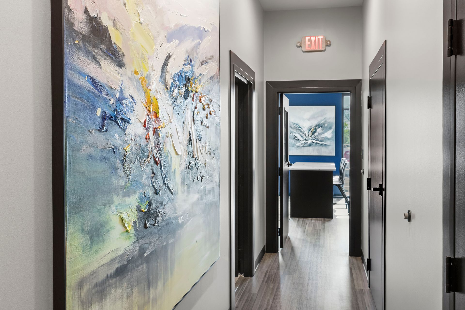 Hallway with abstract art, dark door frames, and exit sign. Light wood flooring leads to a room with a painting.