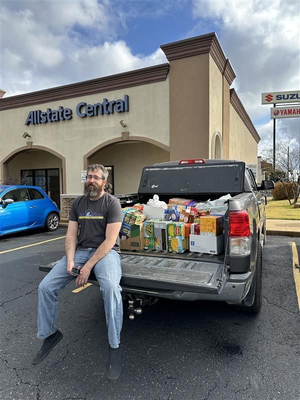 Team member sits on truck bed loaded with food donations outside an office. Sunny day.
