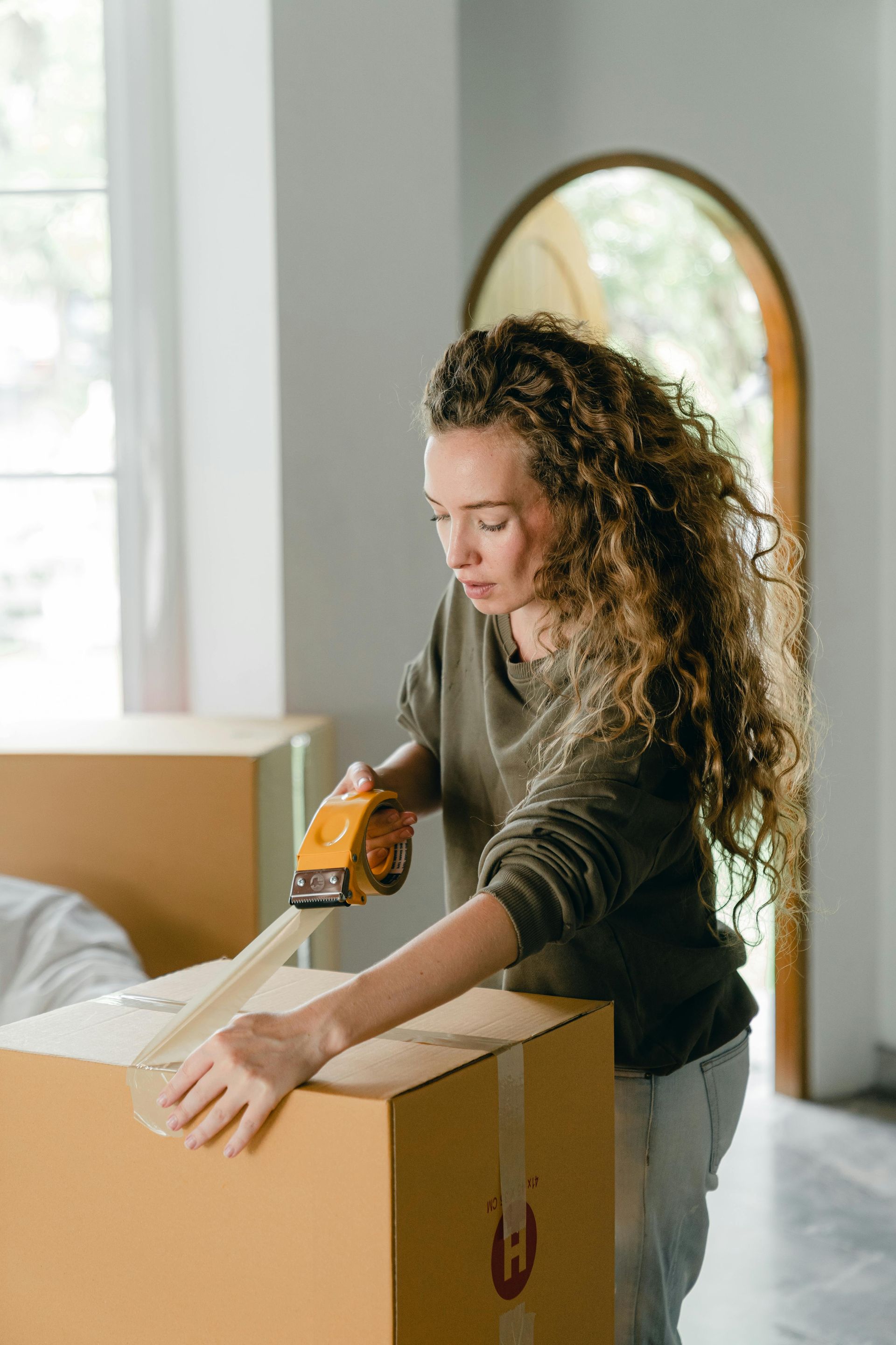Woman taping a cardboard box indoors near a doorway and window; neutral expression.