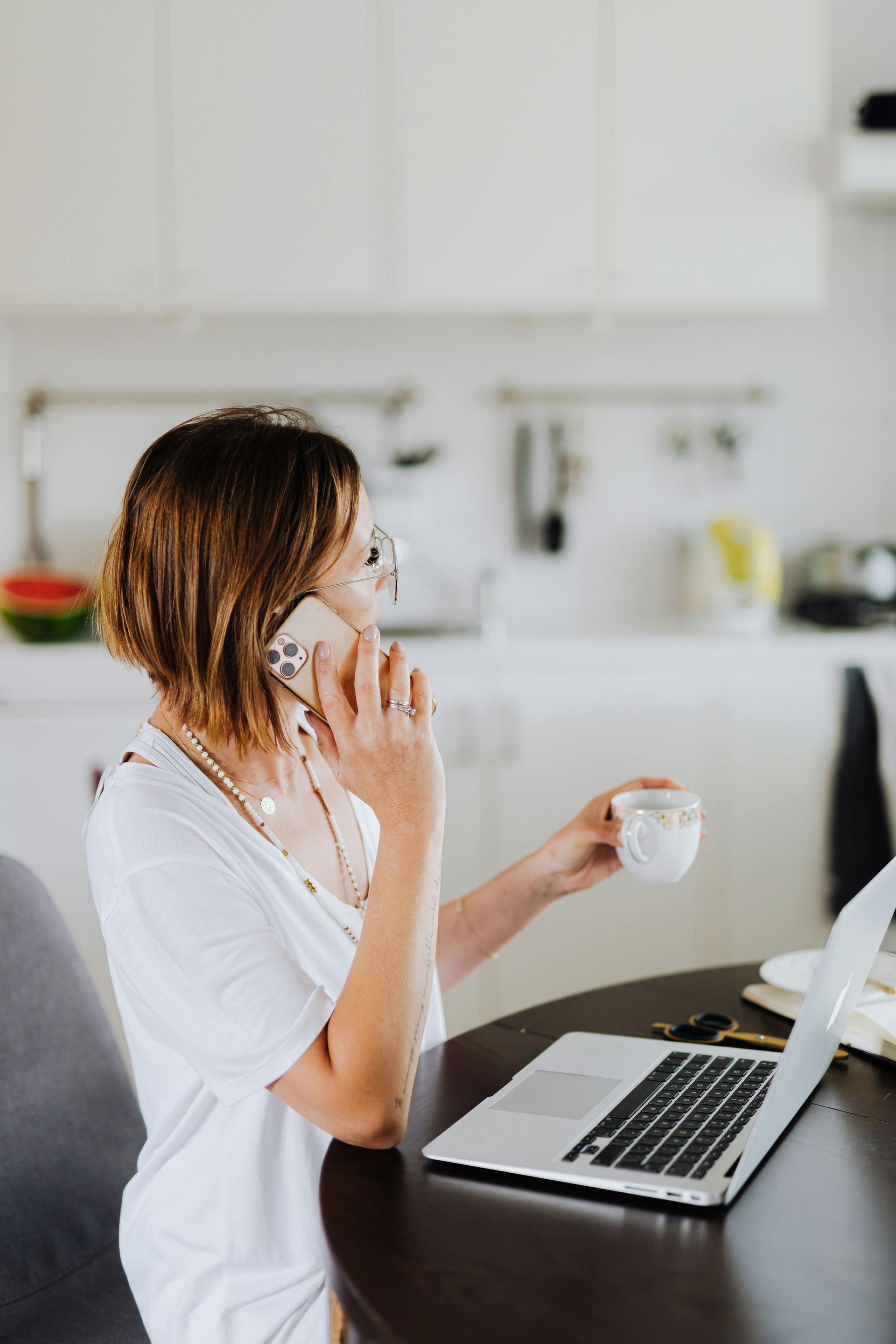 Woman on phone with coffee and laptop in a kitchen setting.