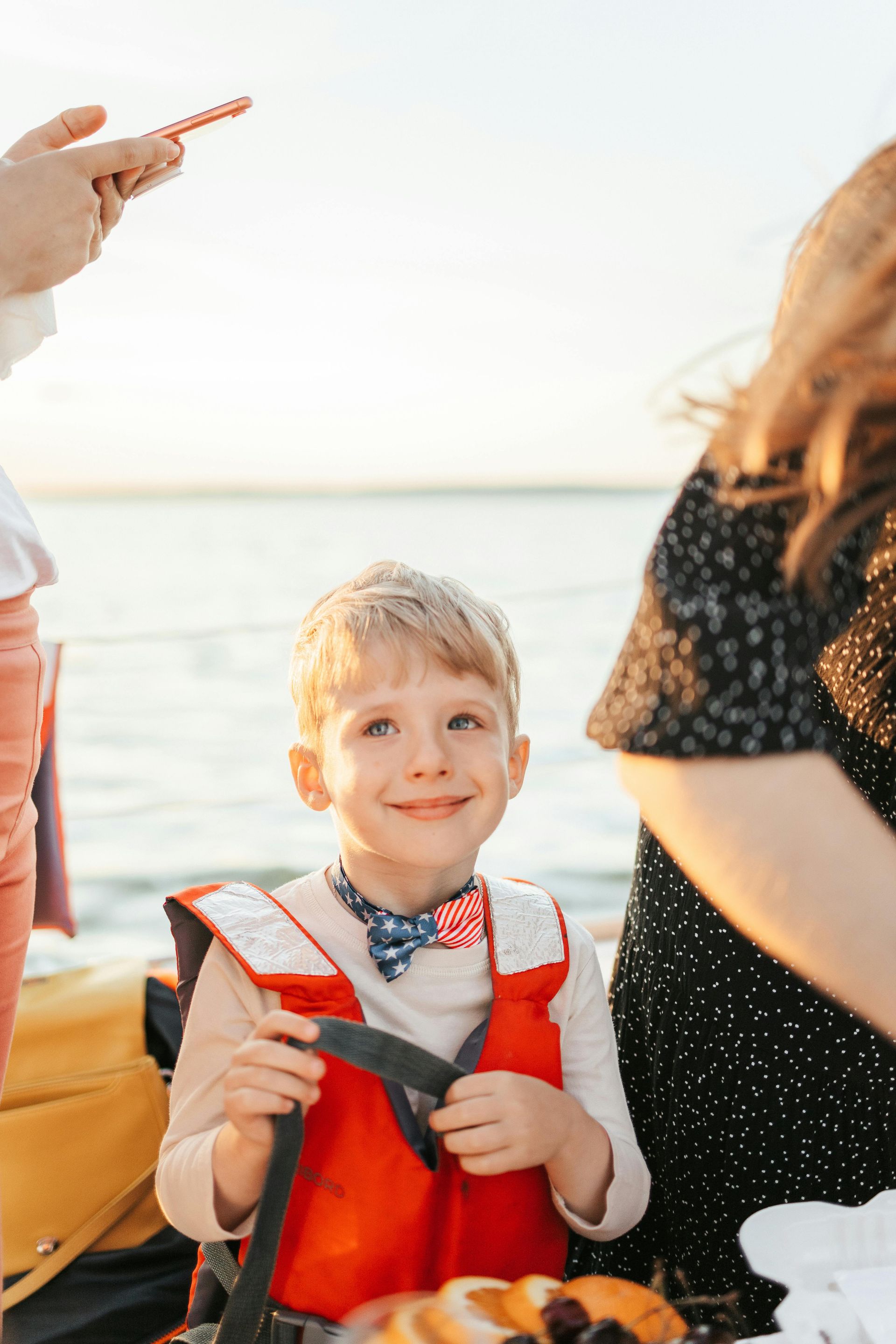 Smiling child wearing a red life vest and a bow tie on a boat; ocean in background.