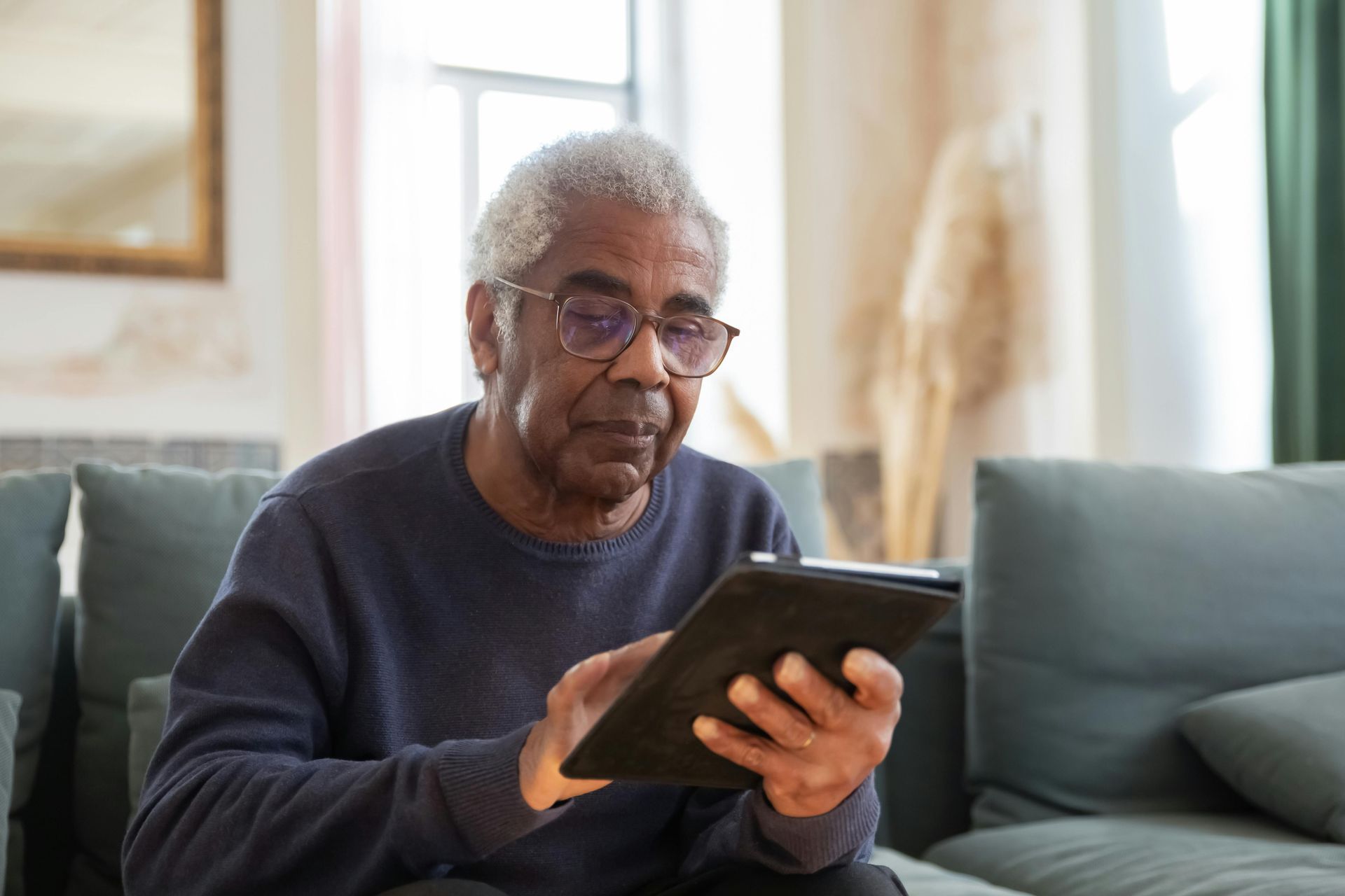 Older person with glasses using a tablet, seated on a couch indoors.