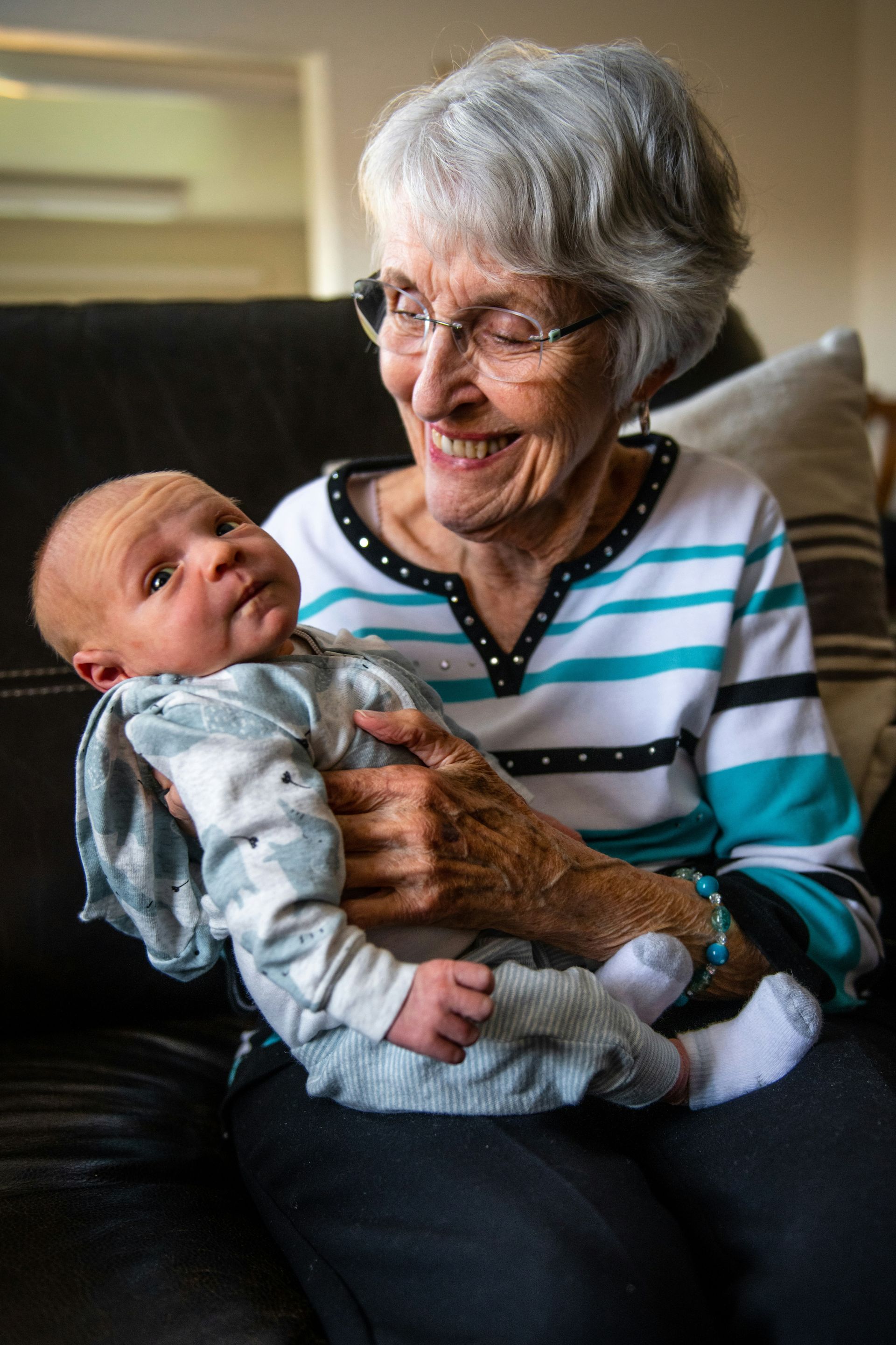 Woman smiling, holding baby. Woman wears glasses, a striped shirt. Baby in a patterned onesie. Indoors, natural light.