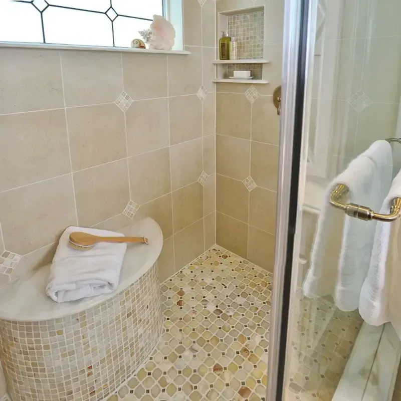 Shower with mosaic tile floor, beige wall tiles, built-in seat with towel, glass door.
