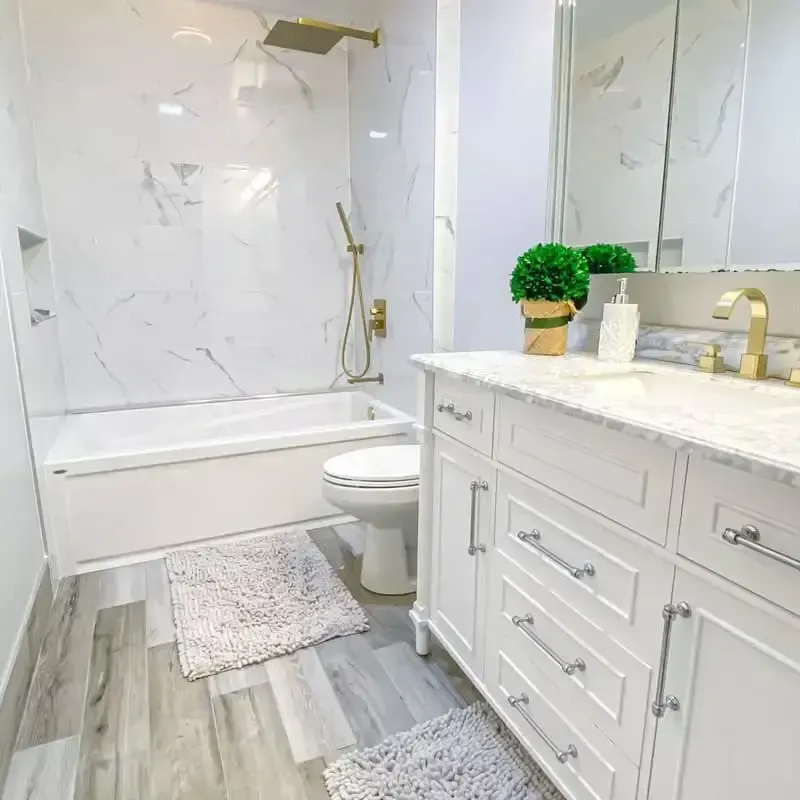 Modern bathroom with white marble shower, vanity, and wood-look flooring. Gold fixtures and a potted plant.