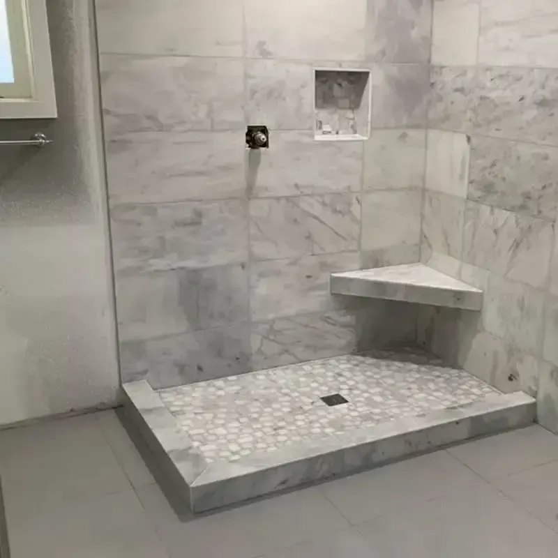 A tiled shower stall with corner seat and recessed shelf. Light gray marble tile covers the walls and floor.