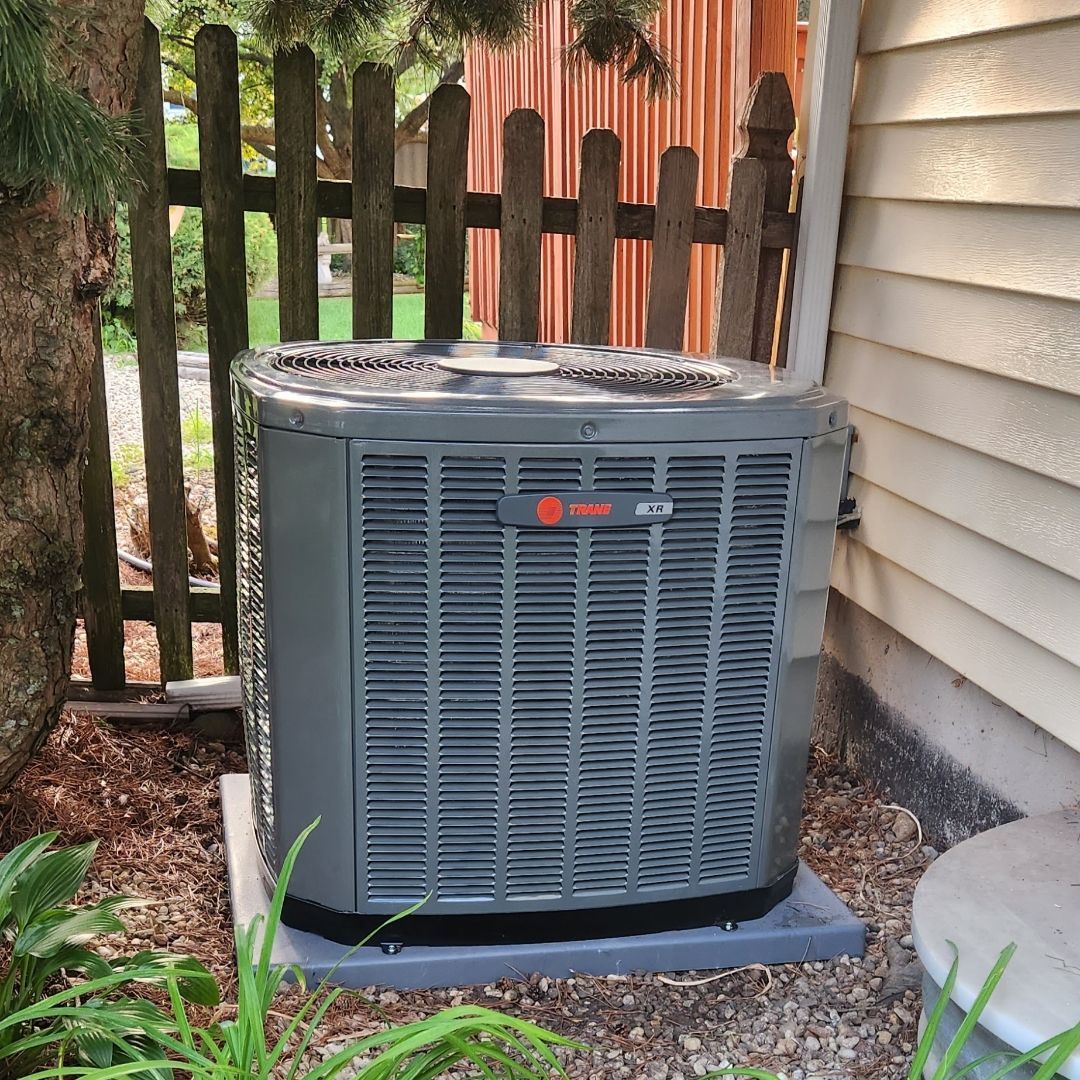 An air conditioner is sitting outside of a house next to a fence.