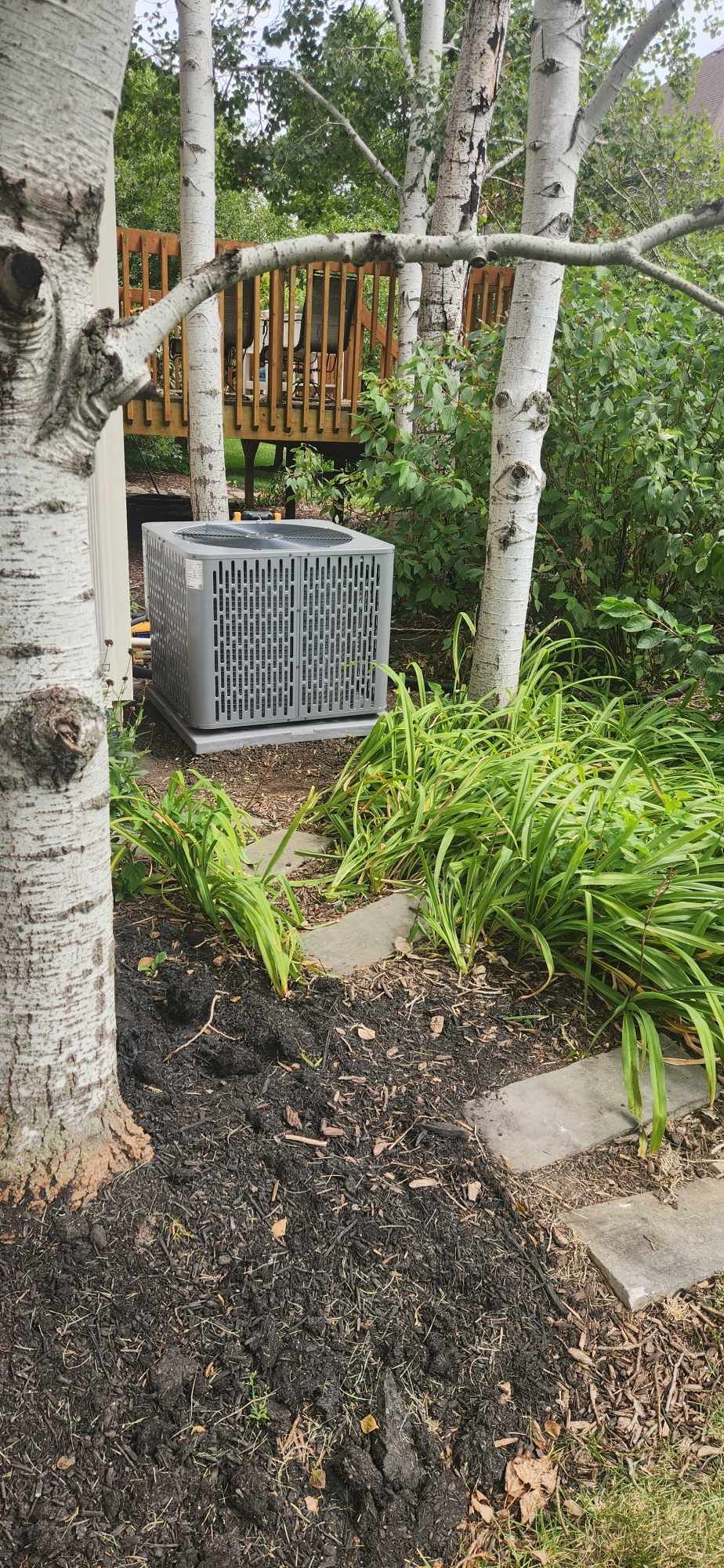 An air conditioner is sitting in the middle of a garden next to a tree.
