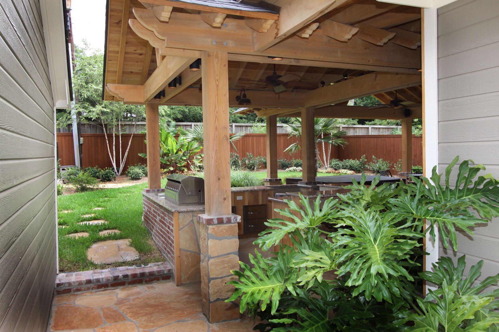 Covered backyard patio with wooden posts, brick base, and lush green plants beside a house