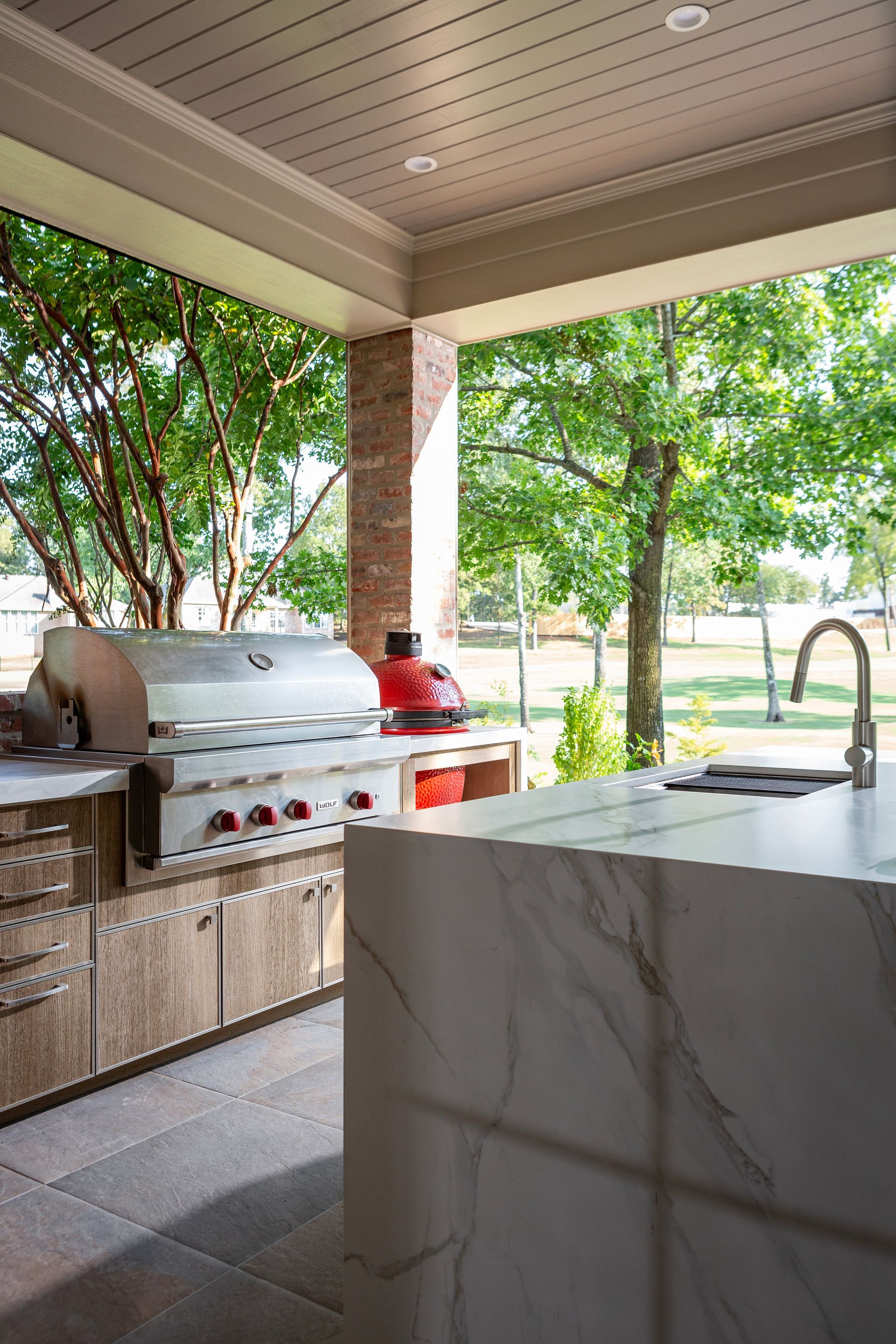 Outdoor kitchen with stainless grill, marble island, and trees beyond a covered patio