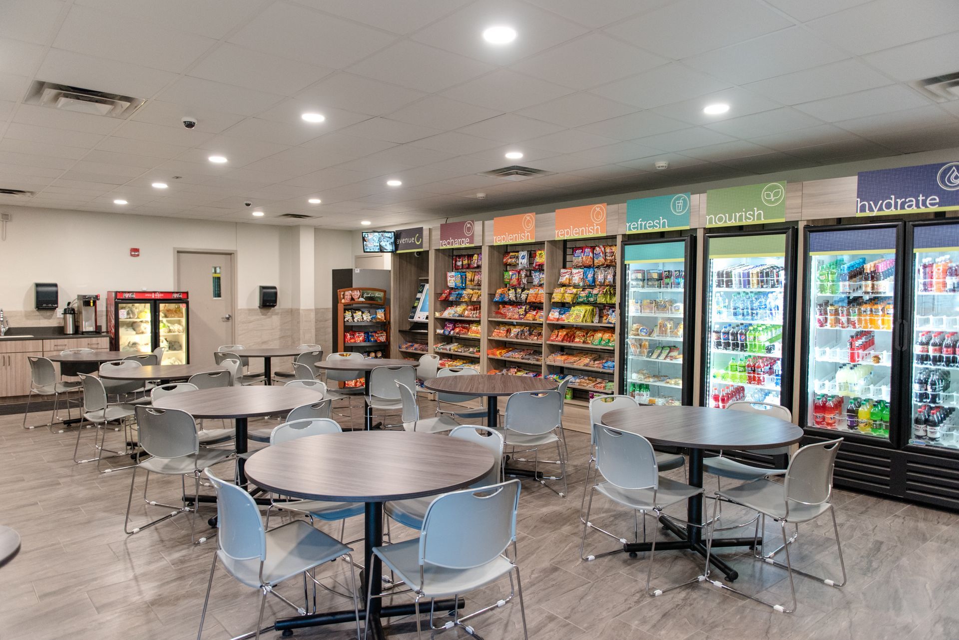Empty cafeteria with round tables, blue chairs, and a row of refrigerated drink coolers.