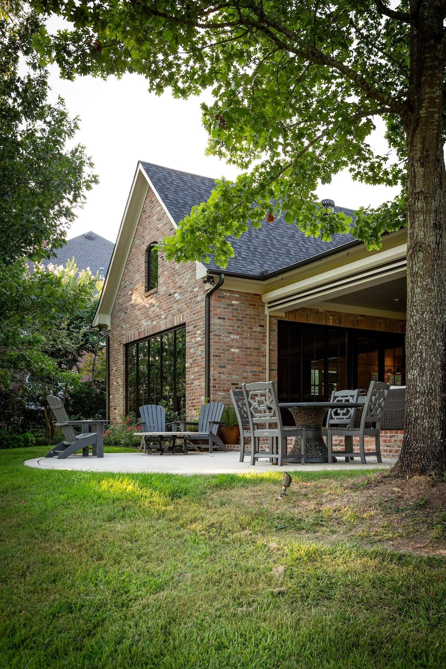 Brick house patio with outdoor dining set, framed by lush green trees and lawn