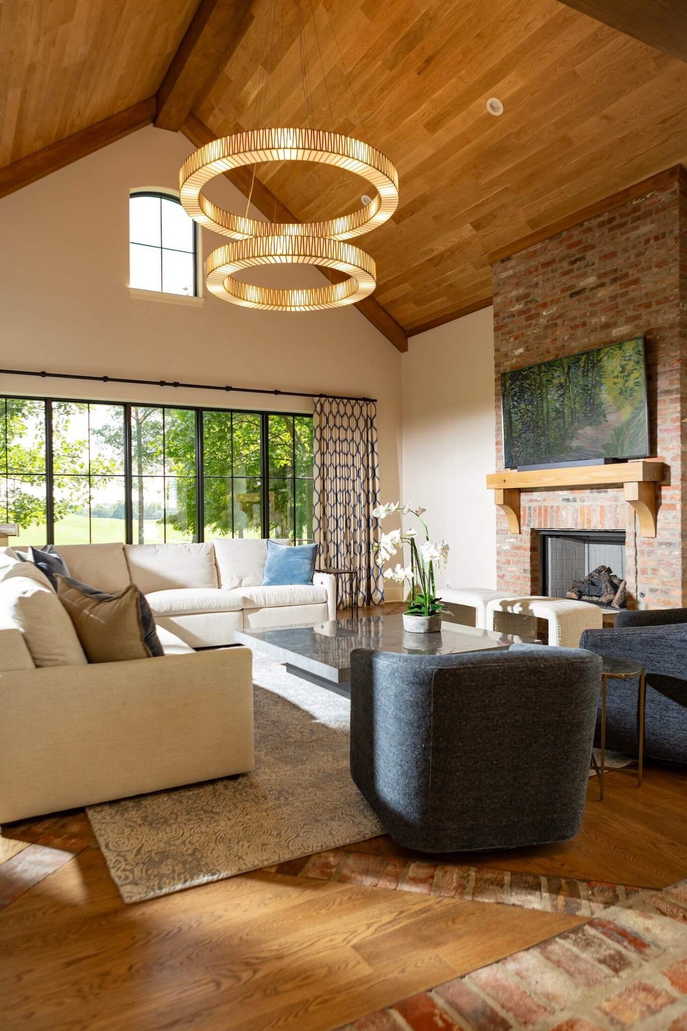 Modern living room with vaulted wood ceiling, brick fireplace, large windows, and beige sectional sofa