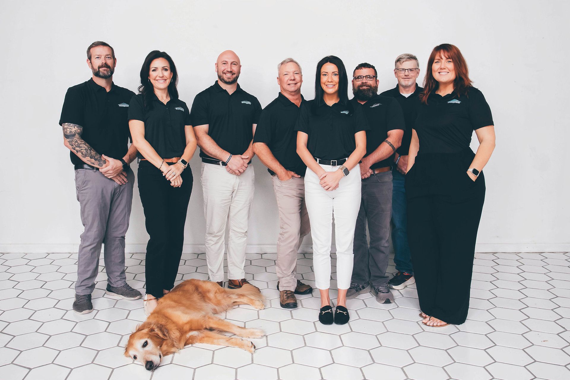 Seven people in black and white stand with a dog on a light brick floor against a white wall.