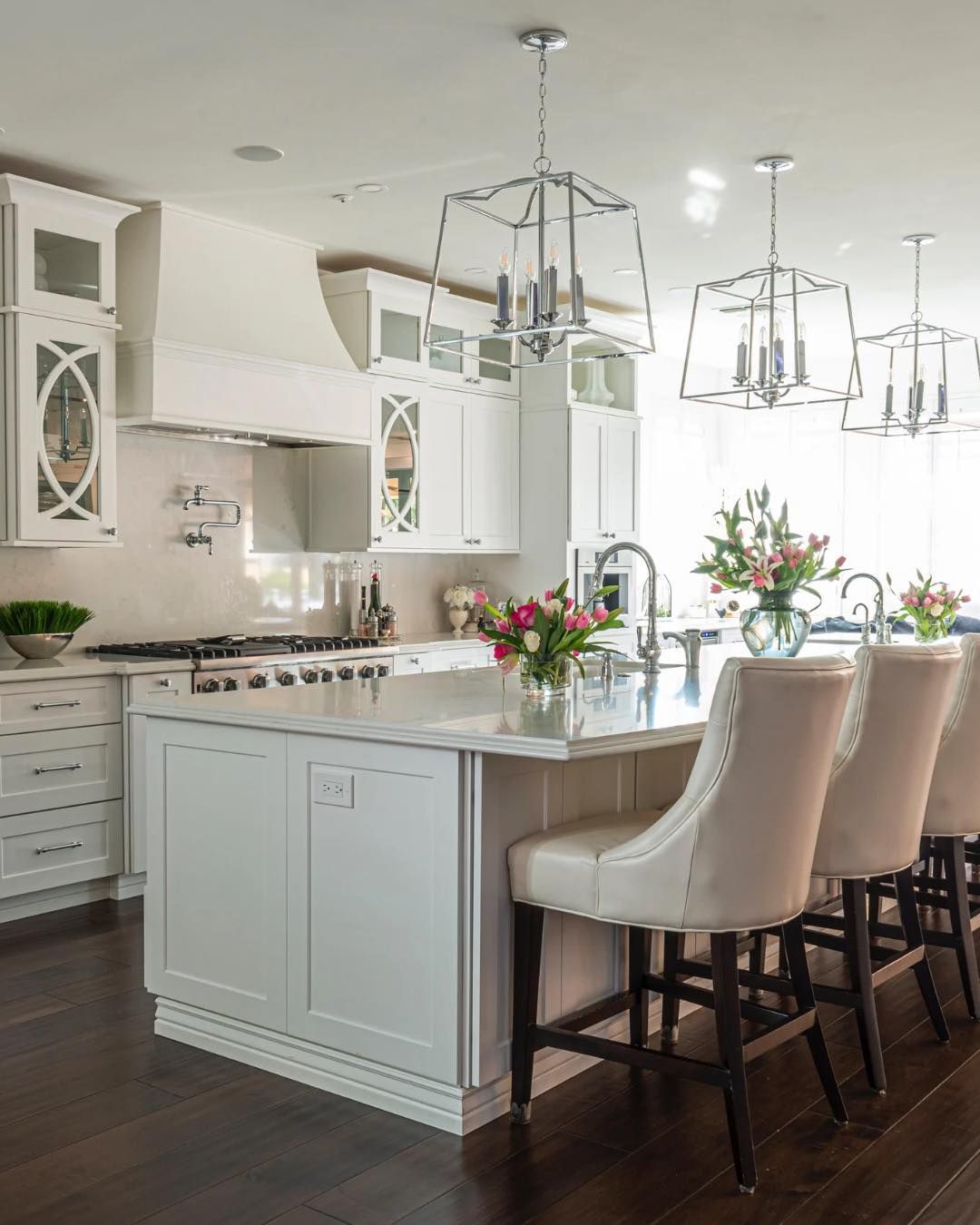 Bright white kitchen with a large island, pendant lights, and upholstered bar stools with flower arrangements.