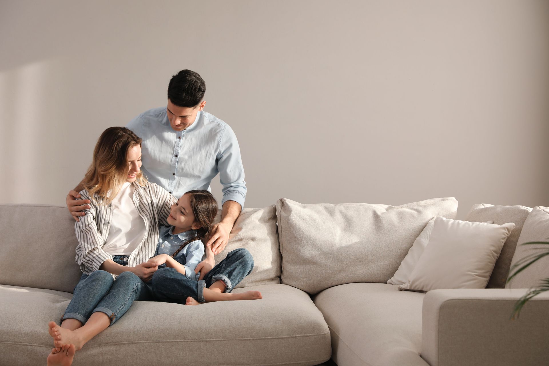 Dreiköpfige Familie auf einem Sofa, der Vater steht über ihnen, warme neutrale Farben.
