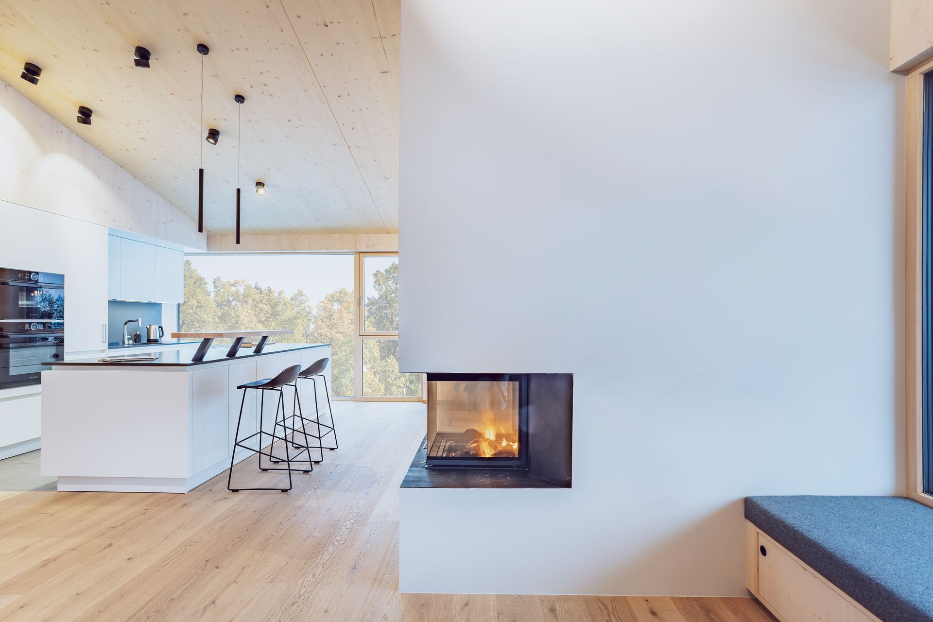 Moderne Küche und Wohnbereich mit weißem Kamin, Holzböden und großen Fenstern mit Blick auf Bäume.