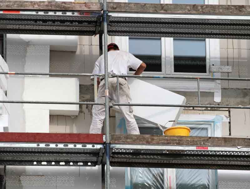 Bauarbeiter installiert mithilfe eines Gerüsts eine Isolierung an der Außenseite eines Gebäudes.