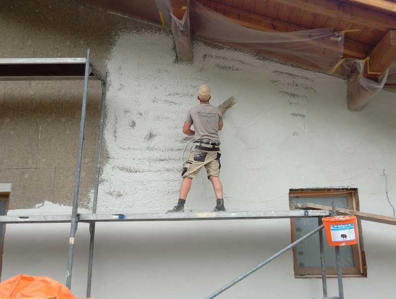Mann auf Gerüst beim Verputzen einer hellen Außenwand. Er trägt Helm und Arbeitskleidung.