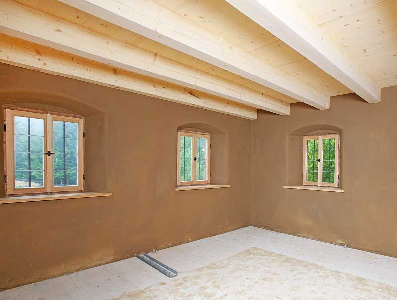 Innenansicht eines Raumes mit erdfarbenen Wänden, drei Fenstern mit Holzrahmen und freiliegenden Holzbalken an der Decke.
