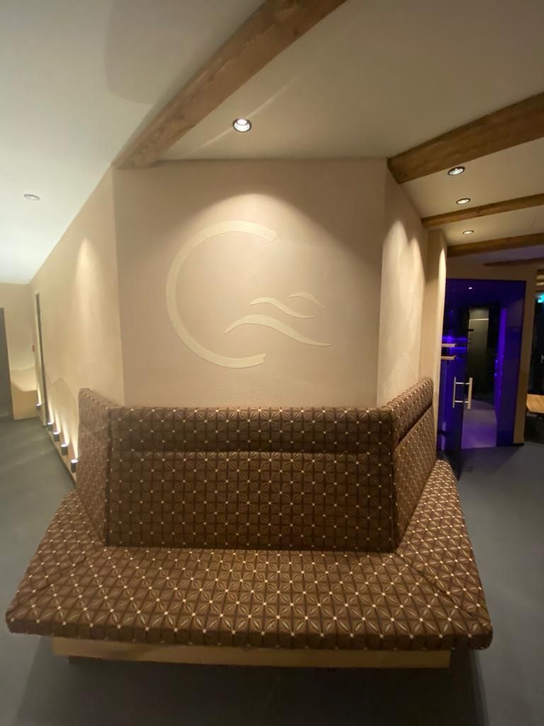 Eine Lounge mit braunem Muster in einem Spa-Ambiente, neutralen Wänden, Holzbalken und einem Logo.