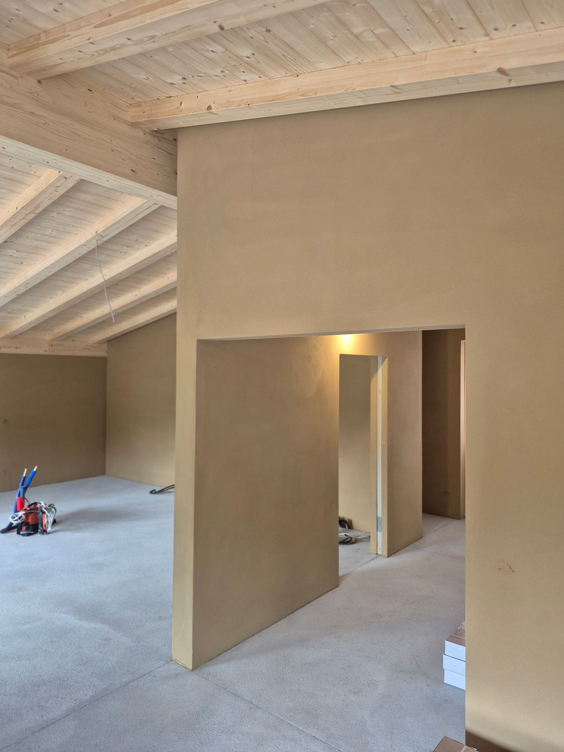Innenansicht eines im Bau befindlichen Raums, hellbraune Wände, helle Holzbalken an der Decke, Betonboden.