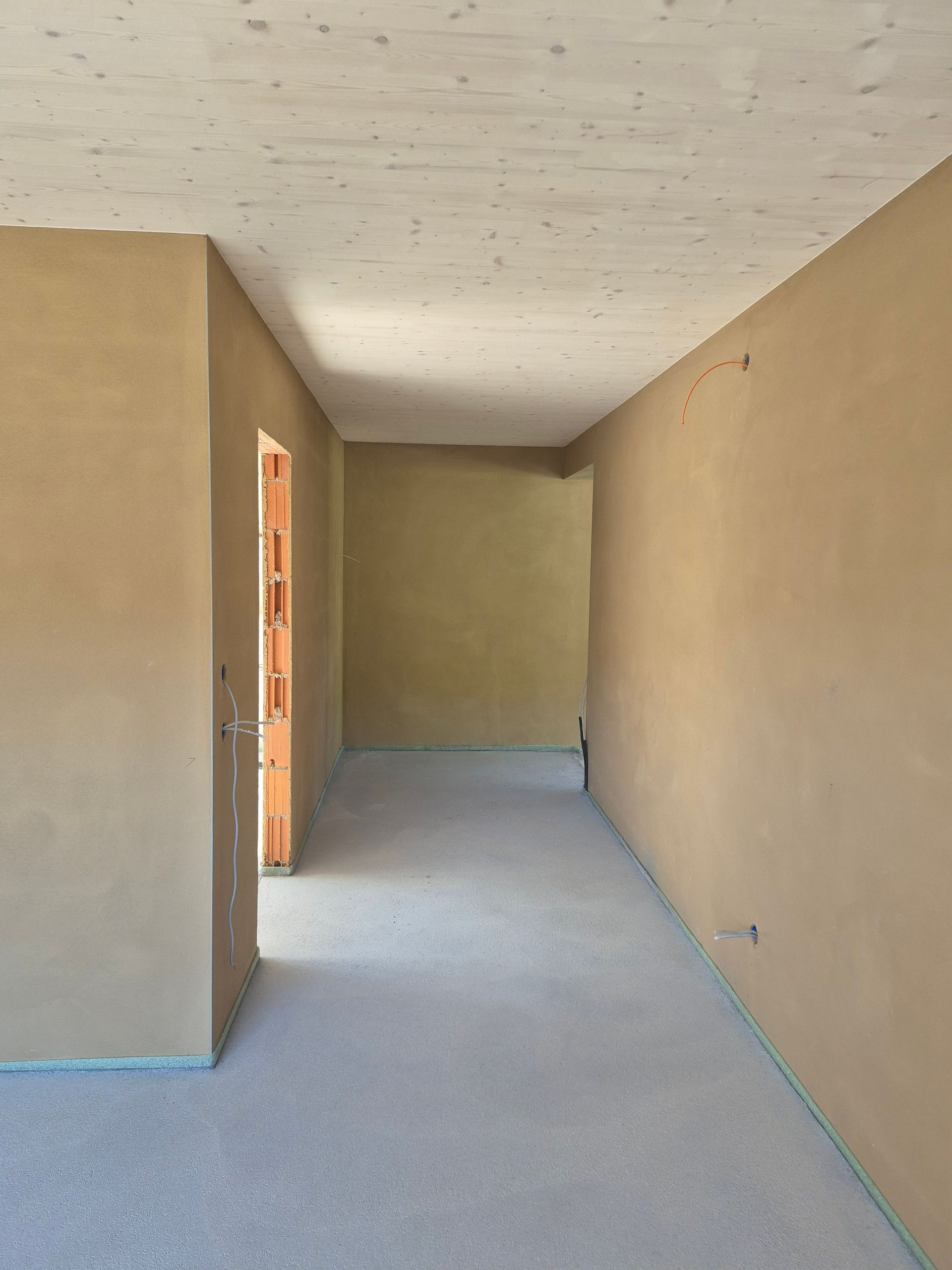 Innenansicht eines im Bau befindlichen Raumes; hellbraun verputzte Wände, offene Tür, Betonboden, Holzdecke.
