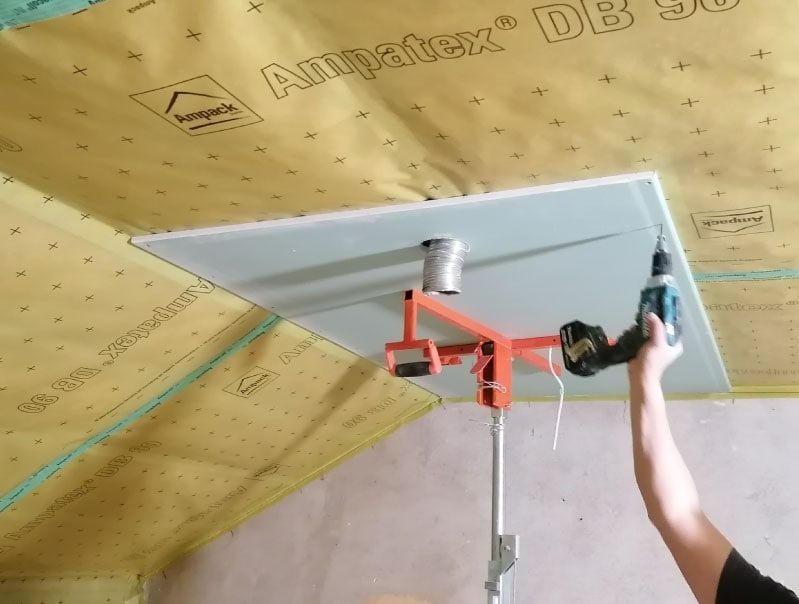 Person schraubt Deckenplatte mit Hebehilfe fest. Gelbe Isolierung, weiße Platte, orangefarbener Lift, Inneneinstellung.