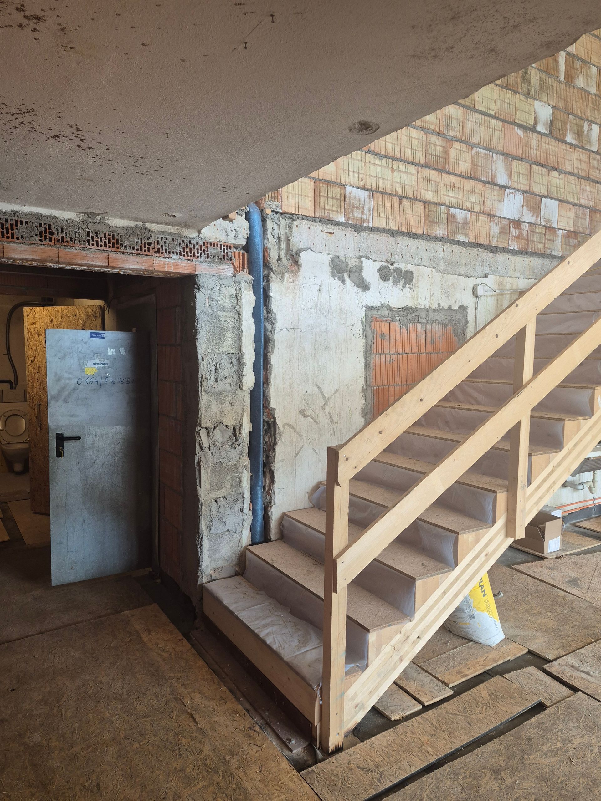 Eine Holztreppe mit Geländer führt in einer Baustelle in der Nähe einer Ziegelmauer und einer offenen Tür nach unten.
