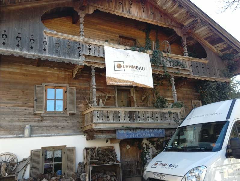 Holzhaus mit Balkon und „LEHMBAU“-Schild; davor parkt ein weißer Lieferwagen.