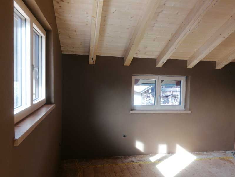 Innenraum mit braunen Wänden, zwei weiß gerahmten Fenstern und freiliegenden Holzbalken an der Decke.