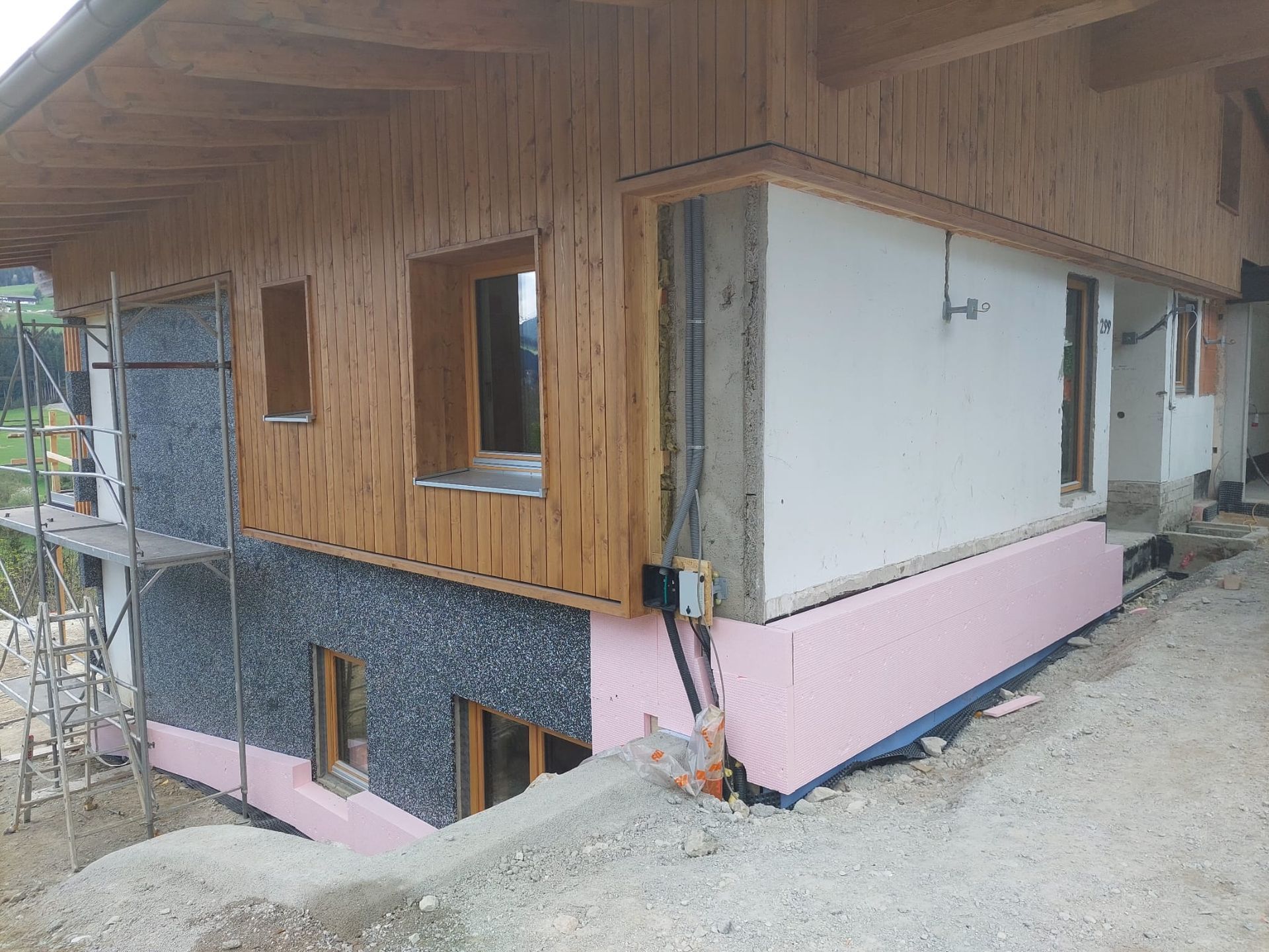 Außenansicht eines im Bau befindlichen Hauses mit Holzverkleidung, Isolierung und freiliegendem Fundament.