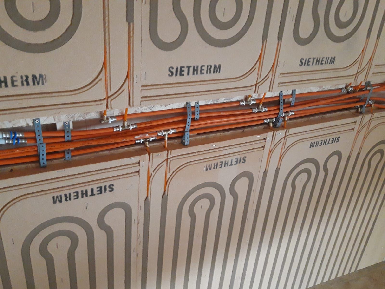 Orangefarbene Rohre und Formstücke, die an eine Fußbodenheizung an einer Wand angeschlossen sind, mit SIETHERM-Etiketten.