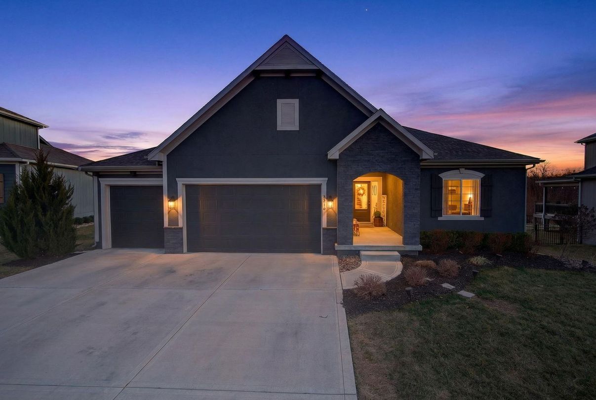 A dark blue suburban house with a two-car garage and a front porch at dusk, under a colorful purple and orange sky.