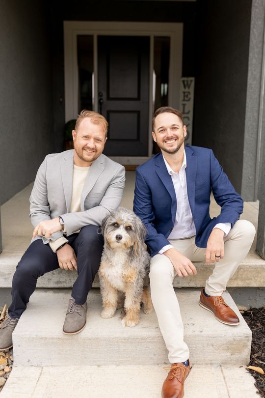 Two men in blazers sit on front steps beside a medium-sized dog in front of a dark front door.