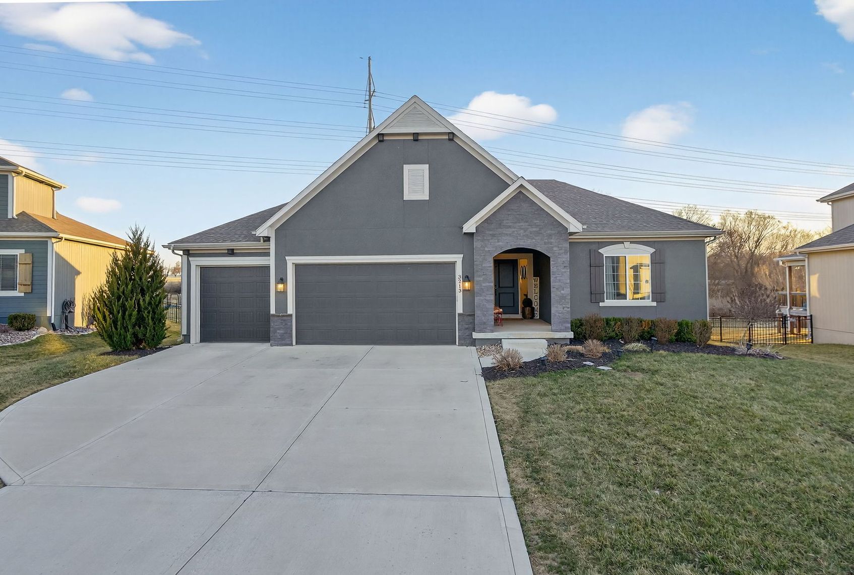 A grey, single-story suburban house with a two-car garage, a concrete driveway, and a small front lawn under a blue sky.