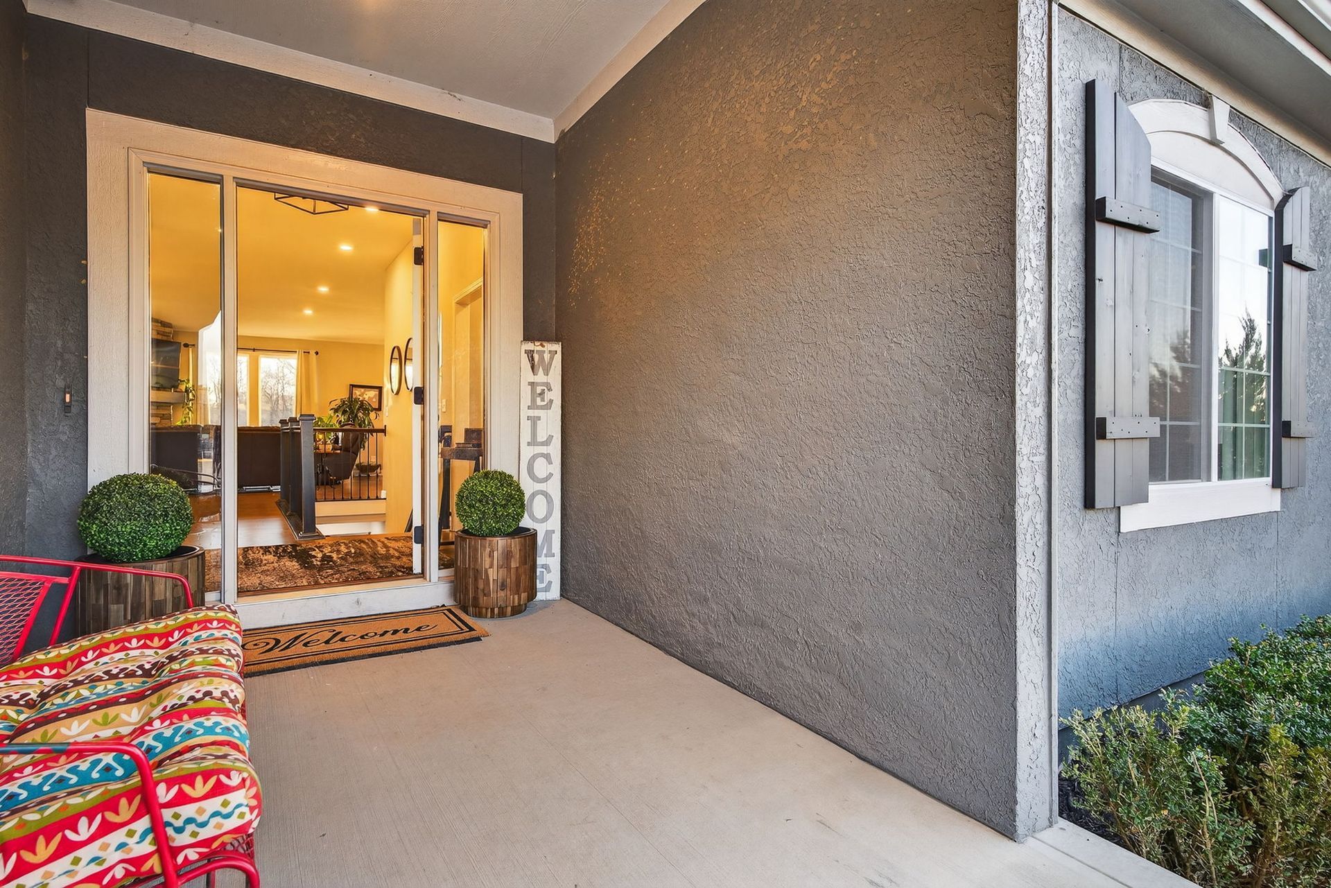 A covered front porch with a patterned chair, a welcome sign, and a view into a bright, open-plan home interior.