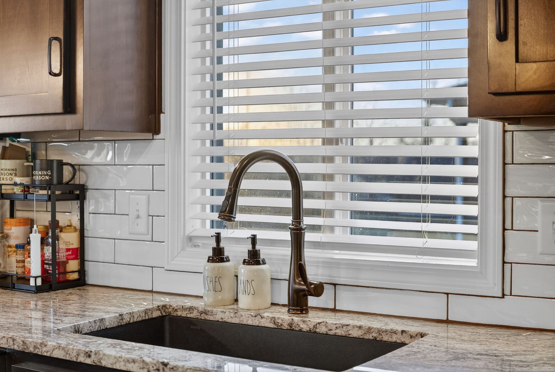 A dark bronze kitchen faucet and two soap dispensers sit on a granite countertop in front of a window with white blinds.