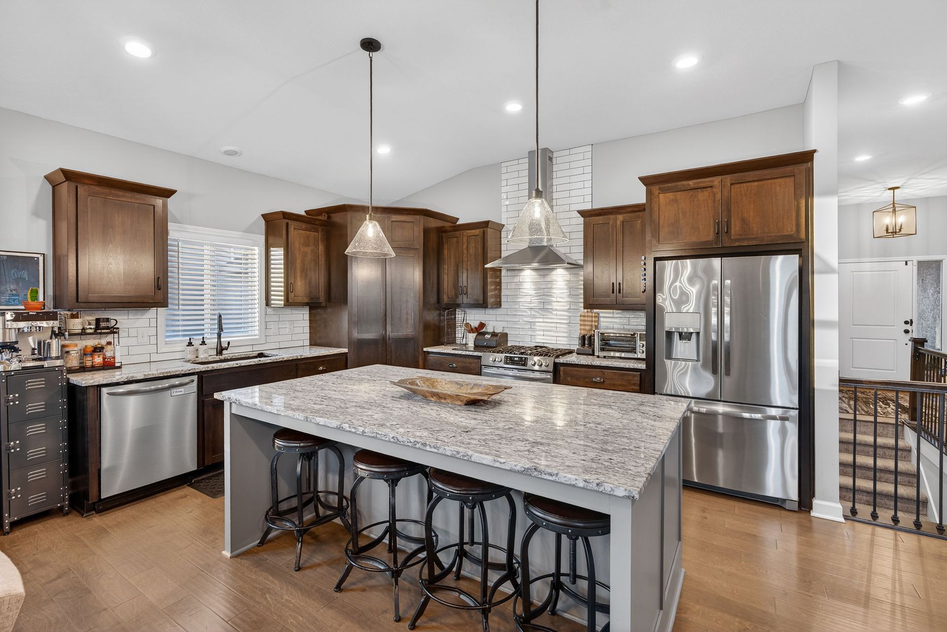 A modern kitchen with brown cabinets, a granite-topped island, stainless steel appliances, and wood floors.