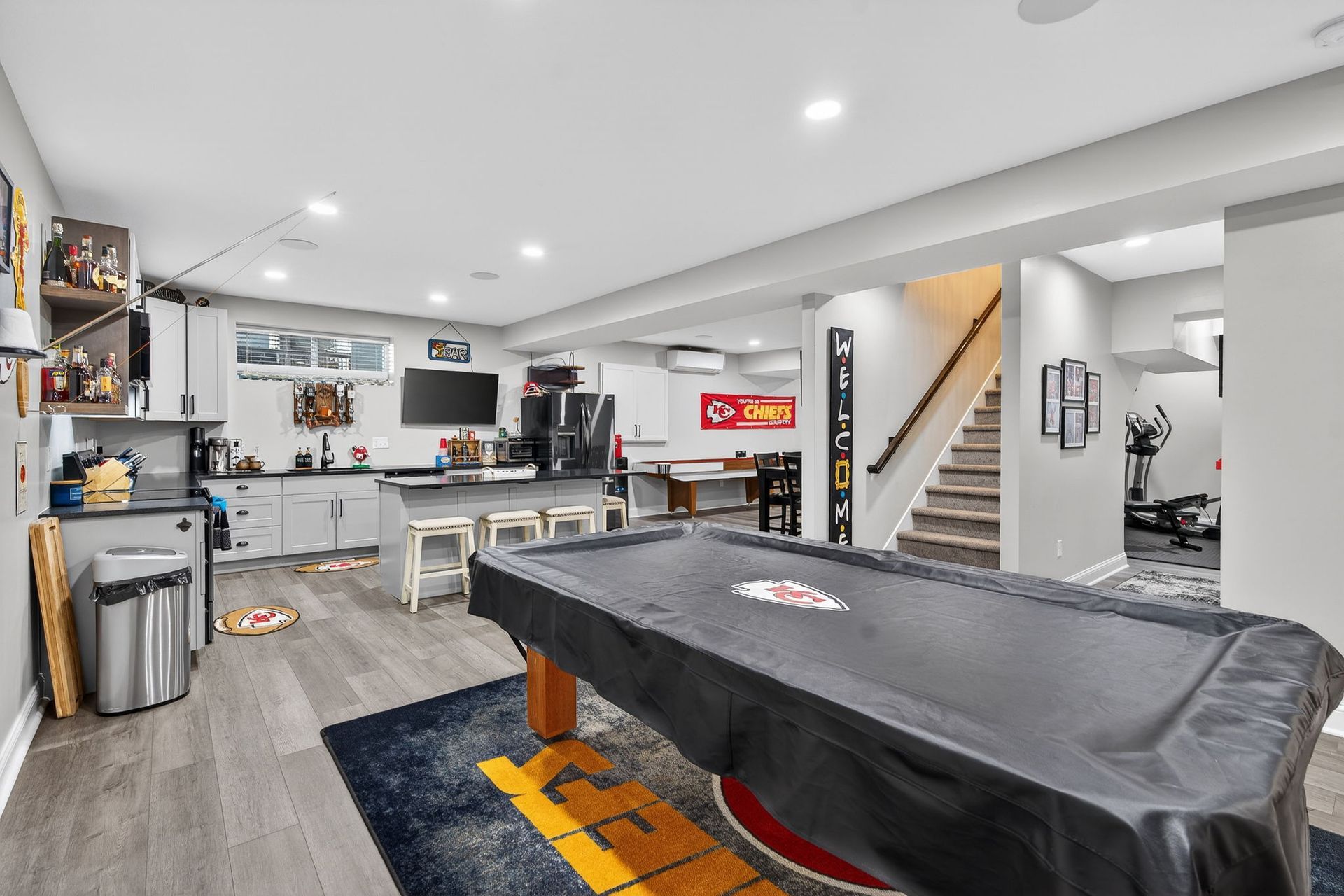 A basement entertainment area with a pool table, kitchenette, bar seating, stairs, and exercise equipment in the back.