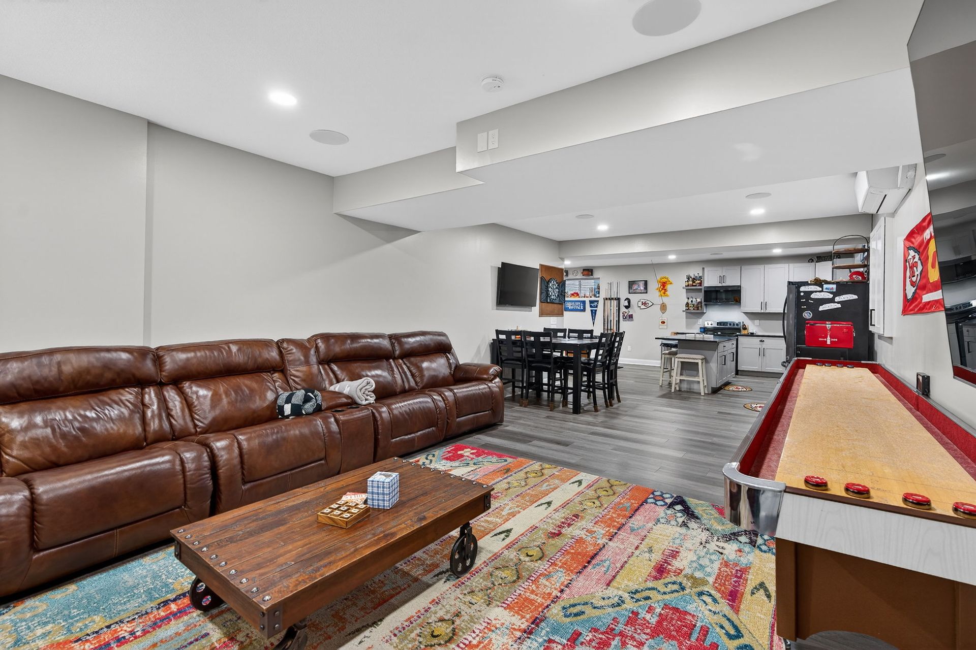 A spacious, open-concept basement with a brown leather sofa, coffee table, patterned rug, dining area, and shuffleboard.