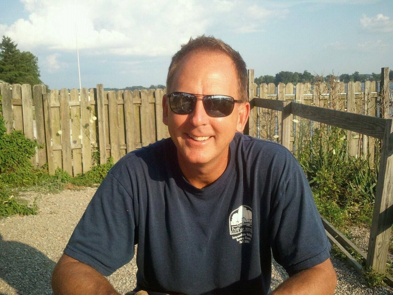 A man wearing sunglasses and a blue shirt is sitting in front of a wooden fence