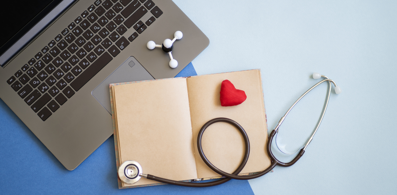 Laptop, open book, stethoscope, pill bottle, heart and syringe on a desk, suggesting online medical care