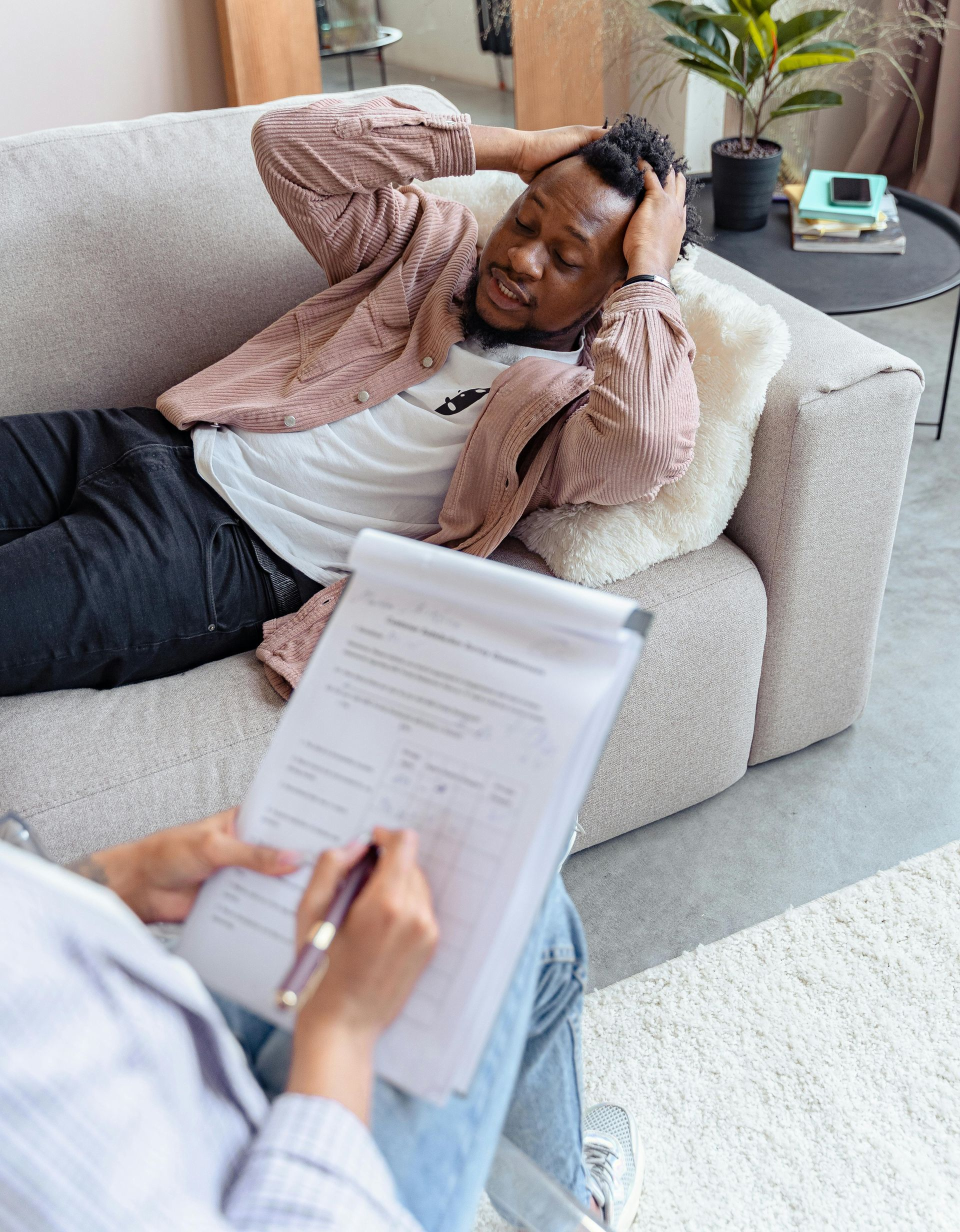 Person resting on a couch while another holds a clipboard and writes in the foreground.