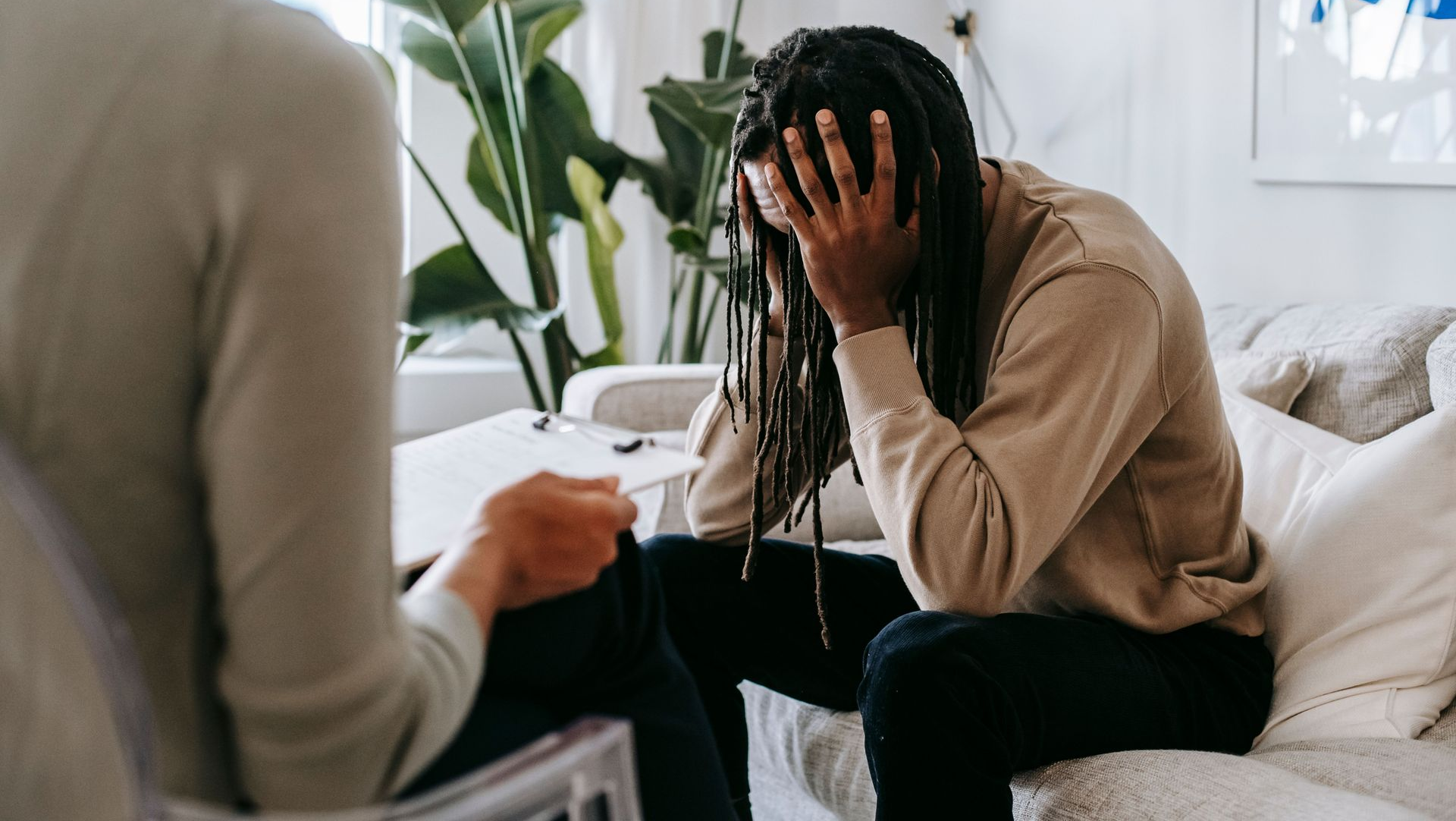 Person sits on a couch, head in hands, talking with a counselor in a bright living room.