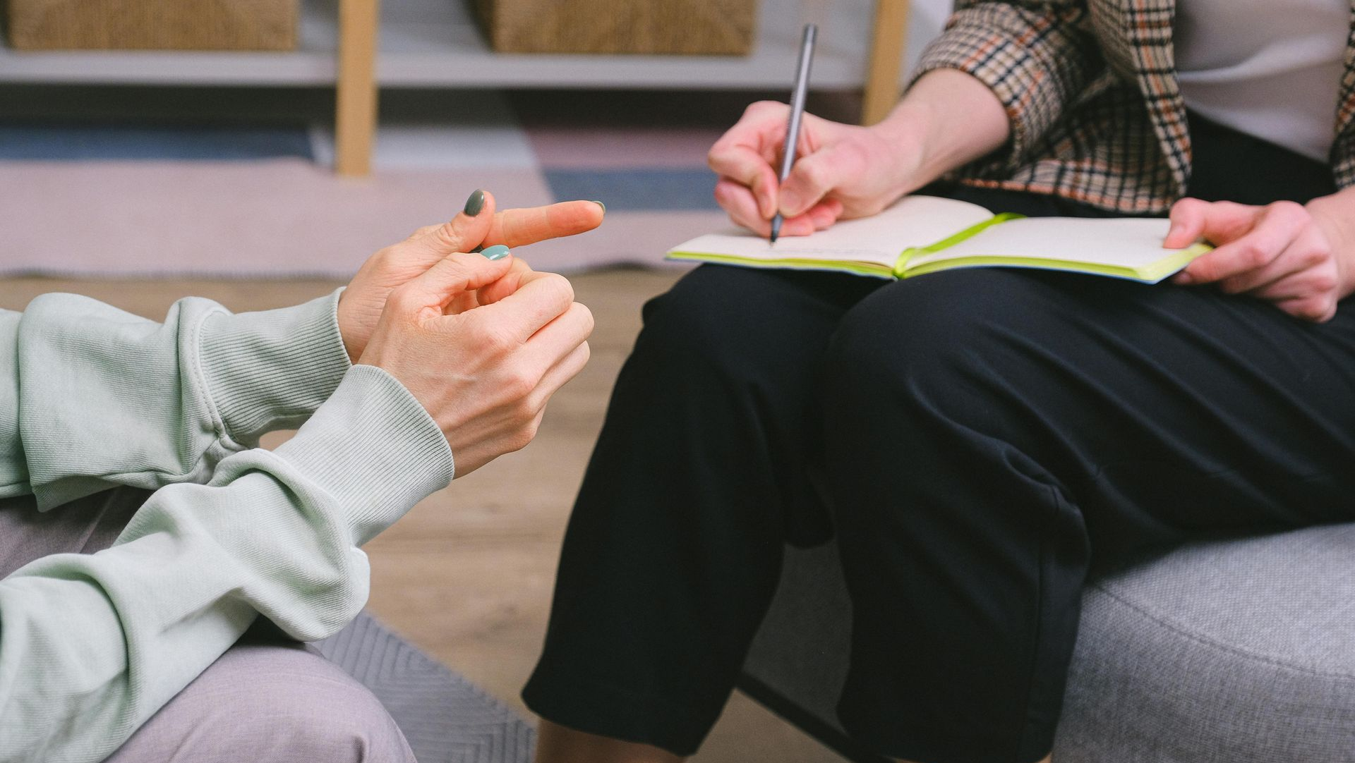 Two people in a counseling session, one taking notes while the other gestures with a pen.