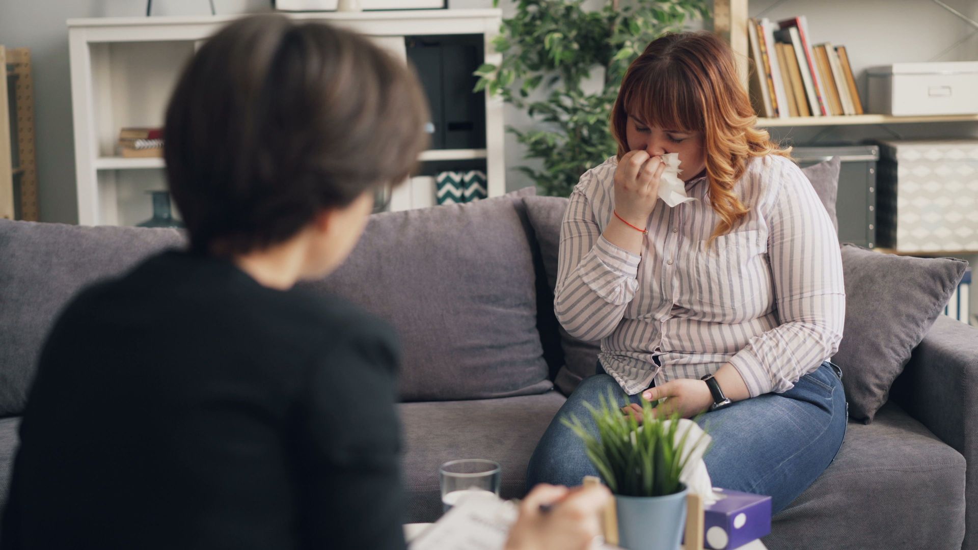 Two people sit on a couch in a living room; one covers her face while the other listens nearby.