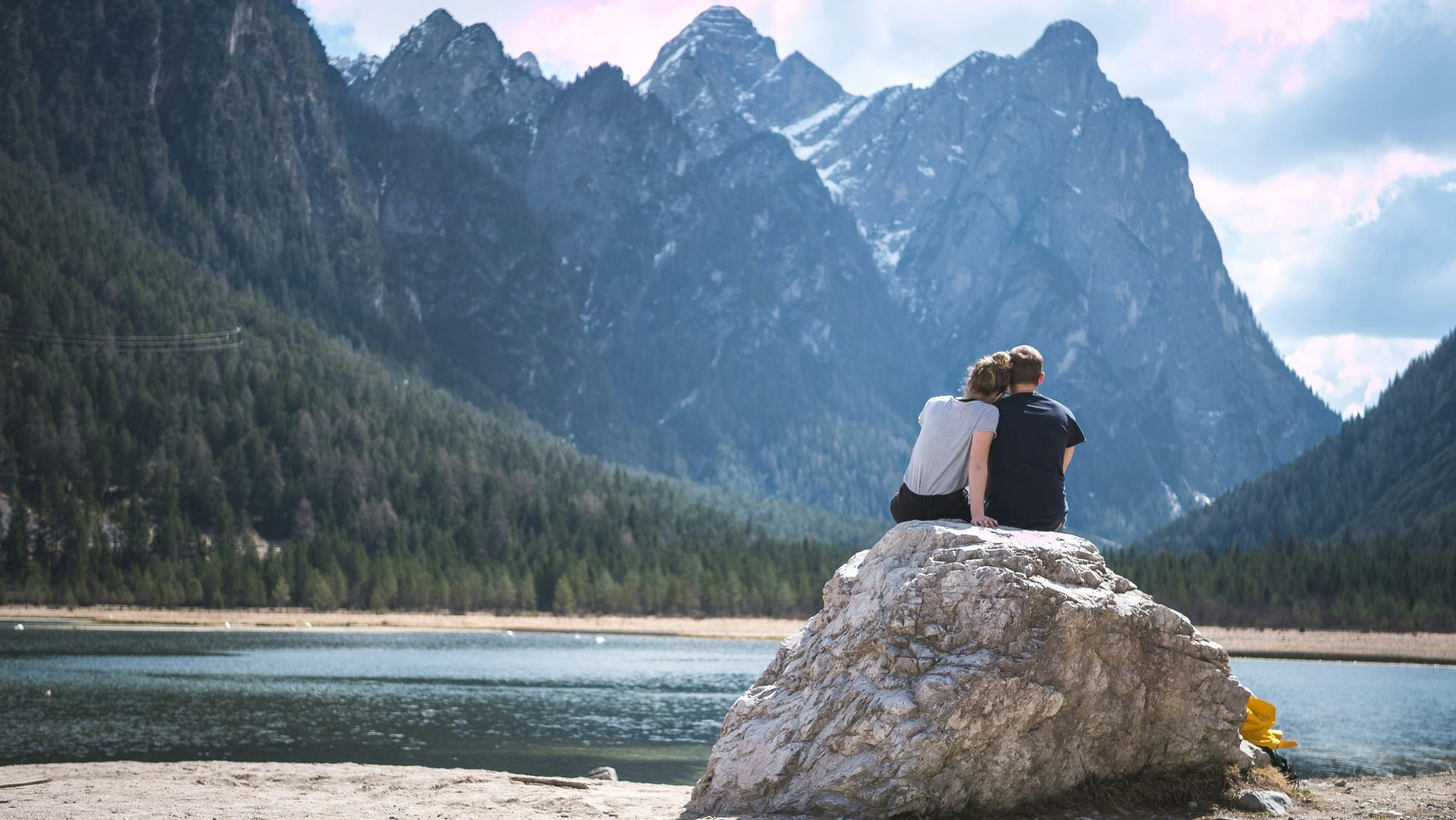 Two people sit on a rock by a turquoise lake with mountains and forest in the background.