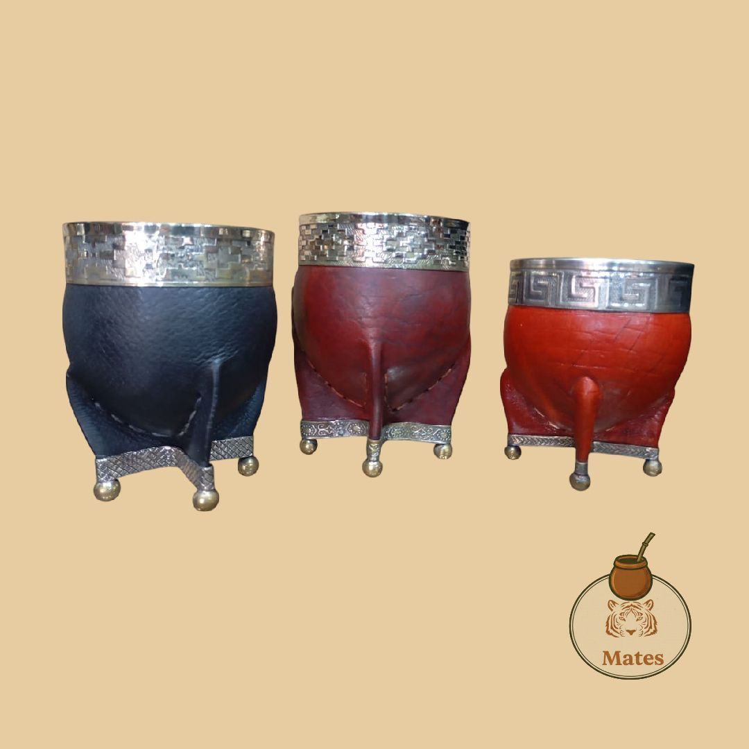 Tres calabazas para mate, negras, granates y rojas, con bordes y patas de metal, sobre un fondo tostado.